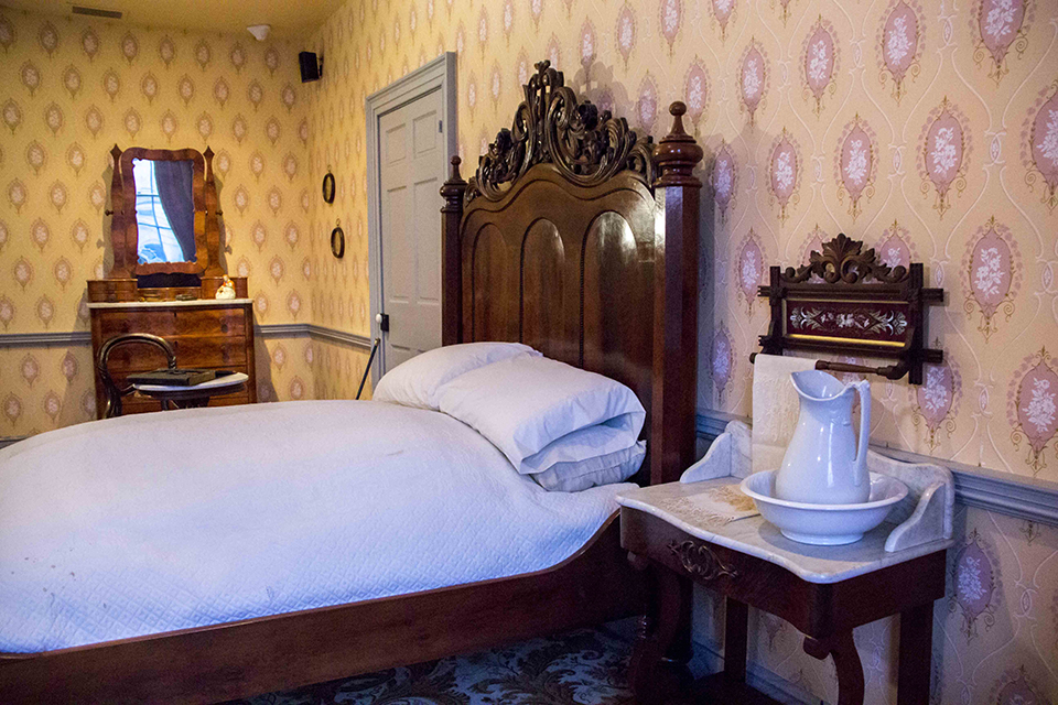 A view from the Lincoln bedroom inside the David Wills House. There is a yellow pattern wallpaper and from left to right: a mirrored dresser, a double bed with ornate headboard, and a night stand with pitcher and basin.