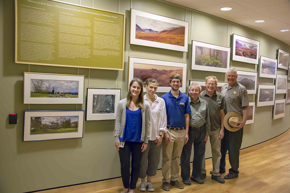 Six people stand in front of the exhibit space where thirteen framed photographs line a green wall.