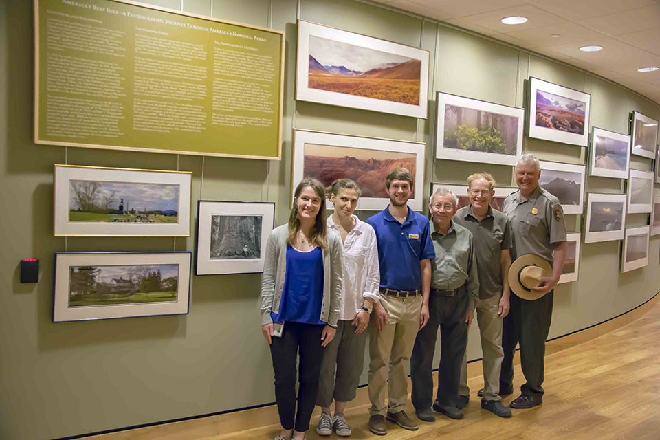 From left to right, Sarah McKenna, NPS intern; Jennifer Weaver, Gettysburg Foundation; David Hazel, NPS intern; Tom and Steve Jorstad, the photographer's sons; and Chris Stein, acting superintendent, Gettysburg NMP and Eisenhower NHS. Six people stand in front of the exhibit space where thirteen framed photographs line a green wall.