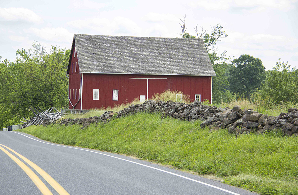 A large red barn sits along the edge of a road, a stone wall runs along the road and a row of trees is behind the barn.
