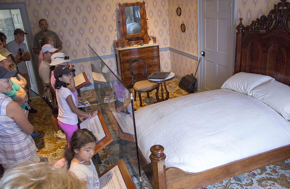 A park ranger talks to a group of visitors in the Lincoln bedroom at the Wills House. On the left are visitors who are looking at the a bed, a dresser with mirror, and a small table and chair.