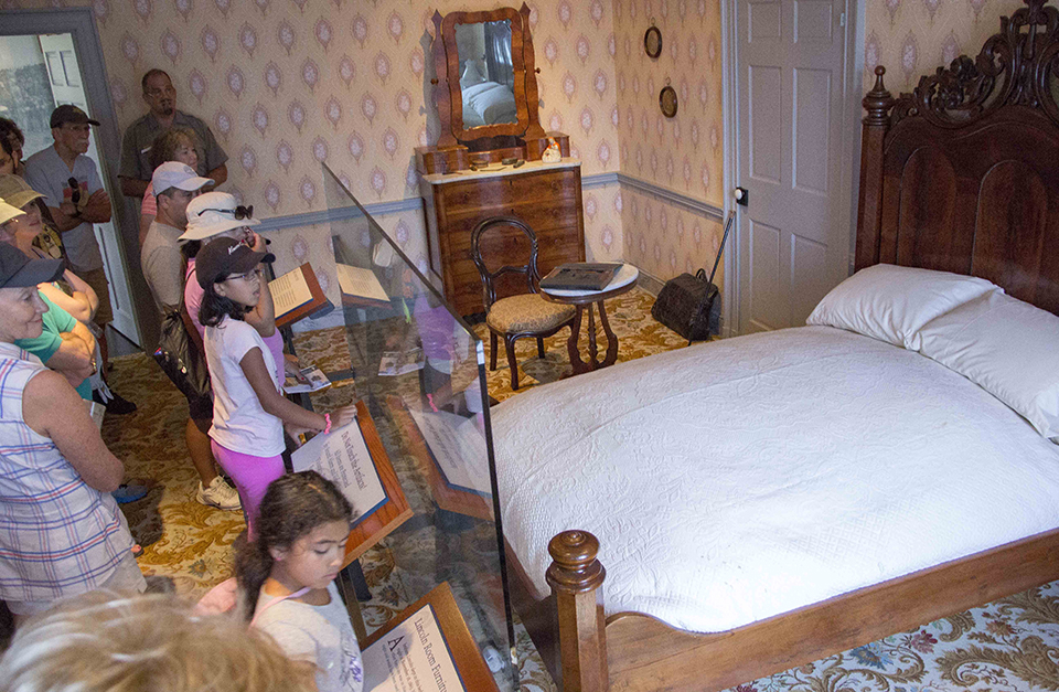 A park ranger talks to a group of visitors in the Lincoln bedroom at the Wills House. A park ranger talks to a group of visitors in the Lincoln bedroom at the Wills House. On the left are visitors who are looking at the a bed, a dresser with mirror, and a small table and chair.