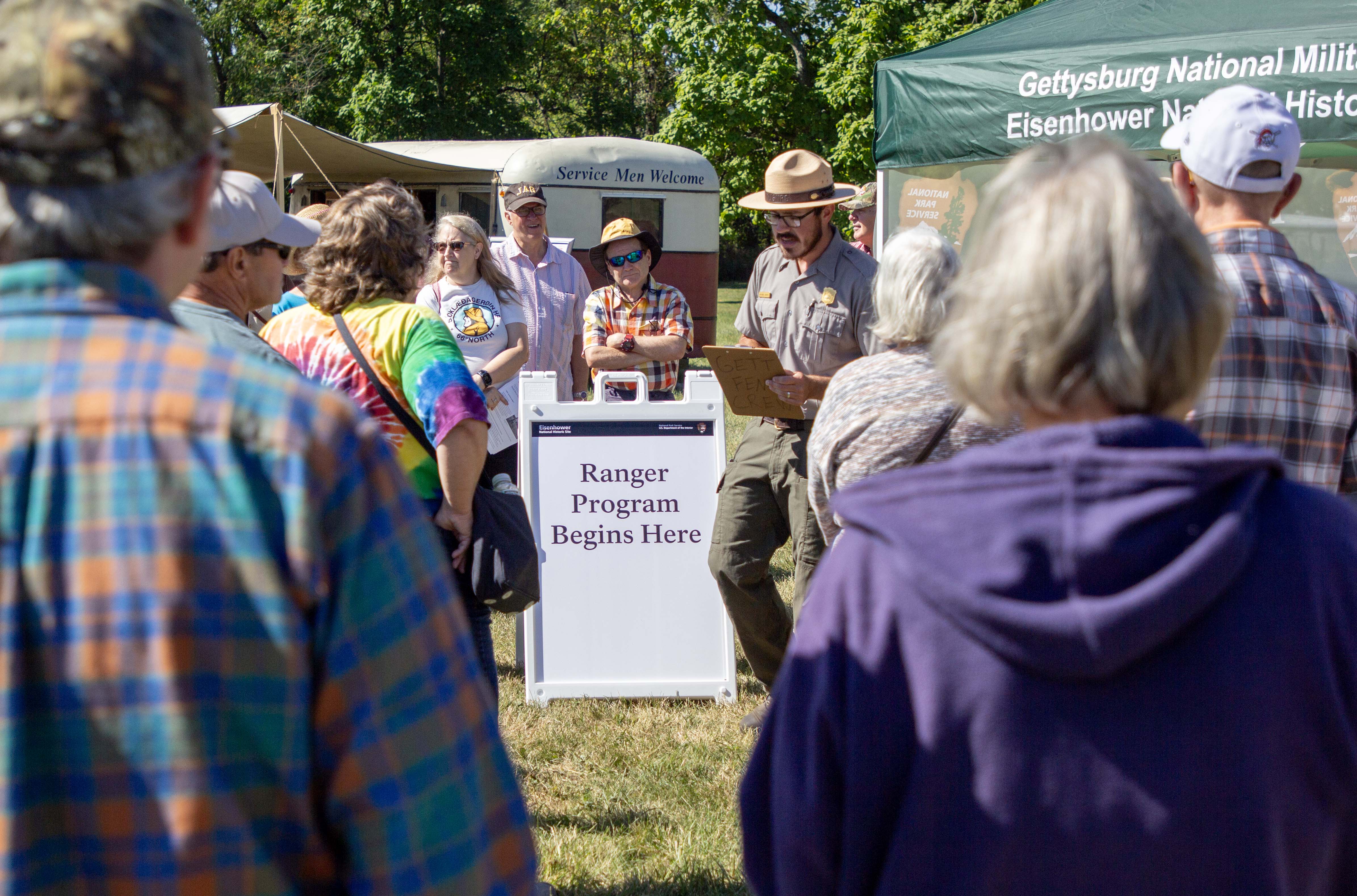 A crowd of visitors gather around a park ranger during the 2023 World War II Weekend held at Eisenhower National Historic Site. Events like World War II Weekend helped support 734 jobs in the local area in 2023. A group of visitors gather around a Park Ranger. In the center is a sign that says Ranger Program Begins Here. Behind the crowd is a red and white trailer that says Service Men Welcome. In the upper right is the top of a green tent.