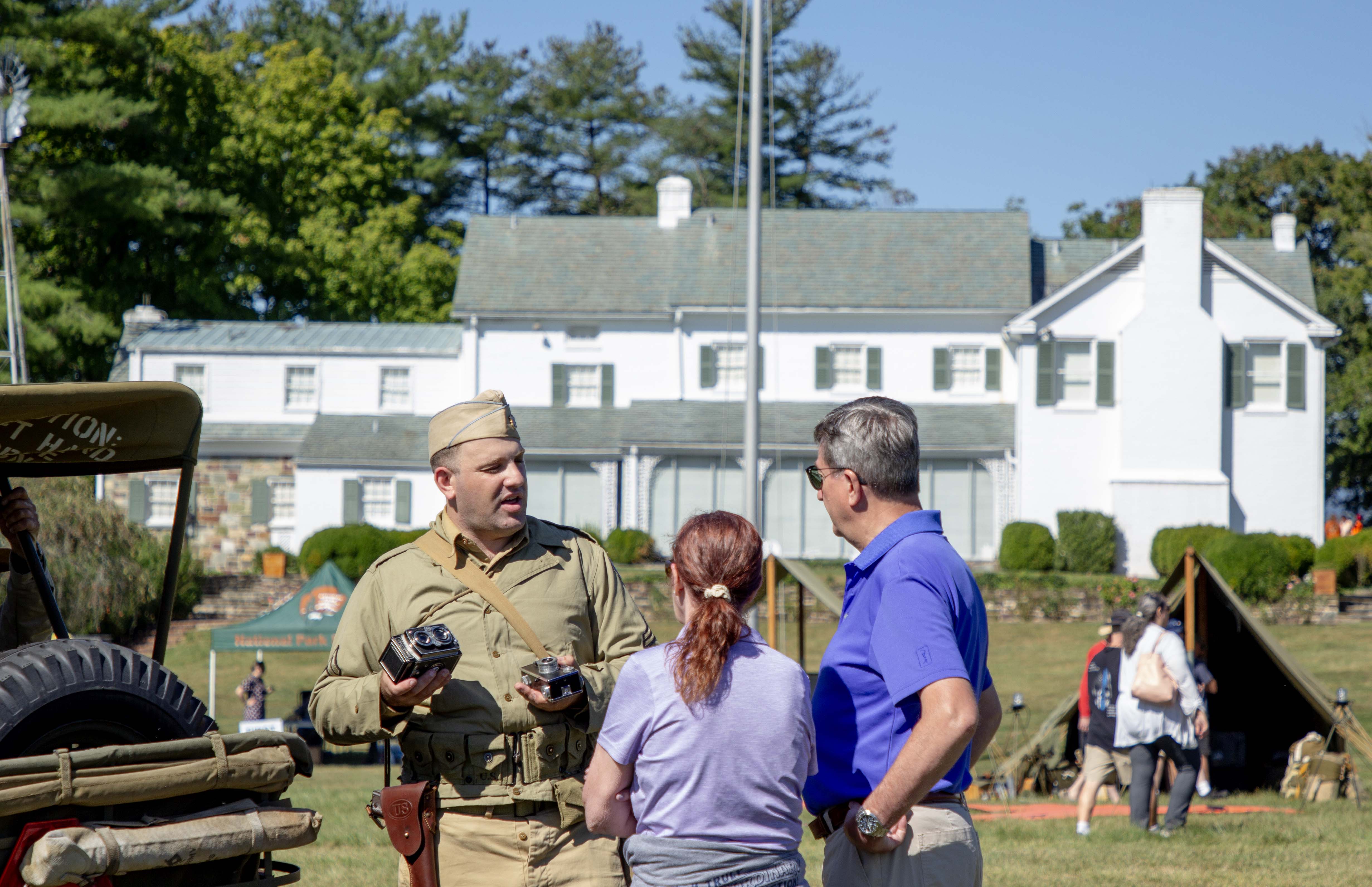 World War II Weekend at Eisenhower National Historic Site. In the foreground is a World War II living historian speaking with two visitors standing next to a Jeep. Behind this group on the right is a khaki colored tent. In the rear is a two-story white house with a green roof and green shutters.