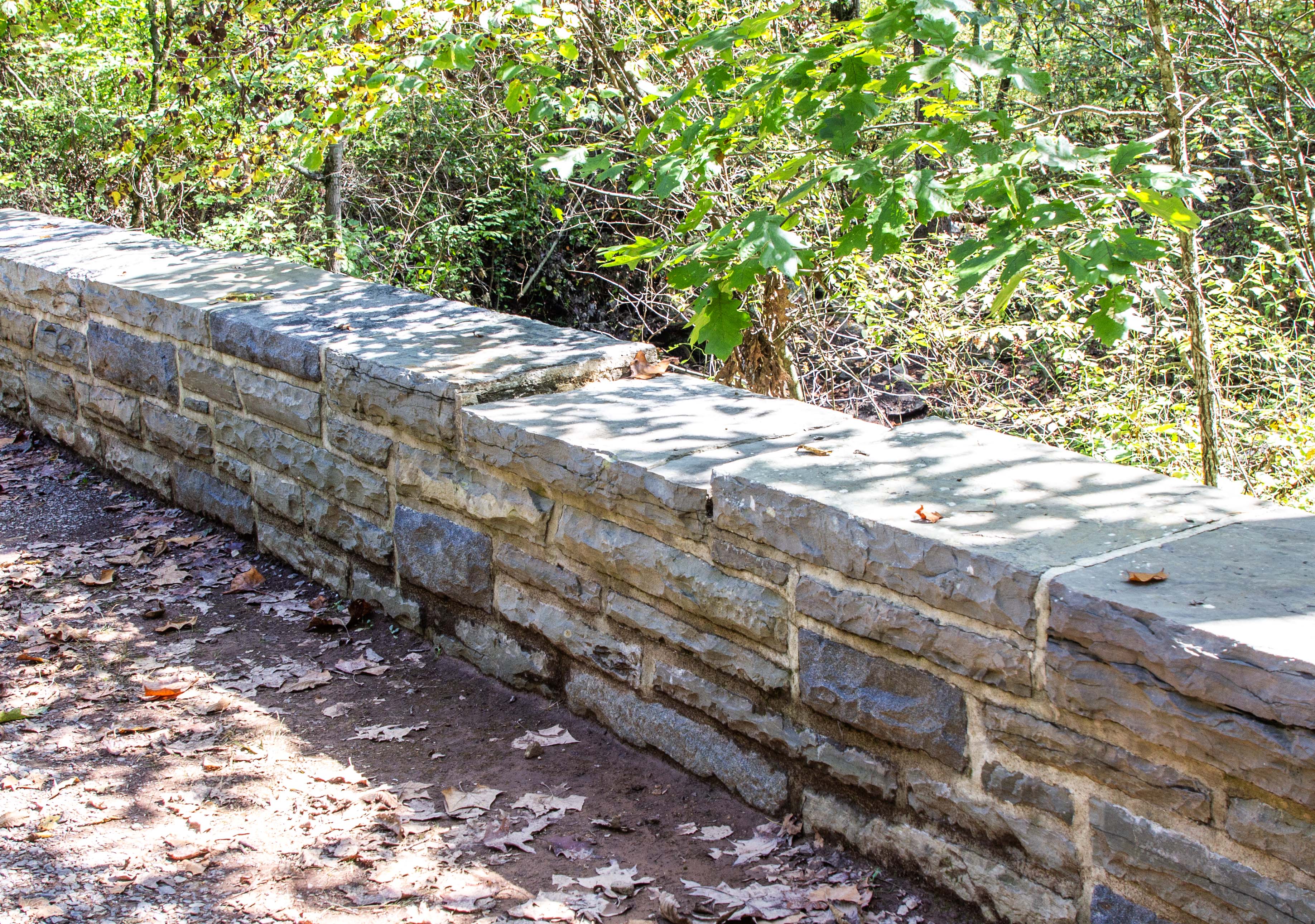 A low stone wall has one of the cap stones missing. To the left is a packed dirt surface. To the right it's heavily wooded.