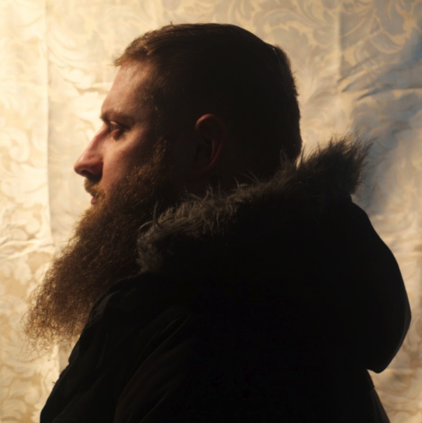 Joshua Osburg Self portrait of Joshua Osburg from his left side. He has a long beard and is wearing a dark coat with a fur lined hood.