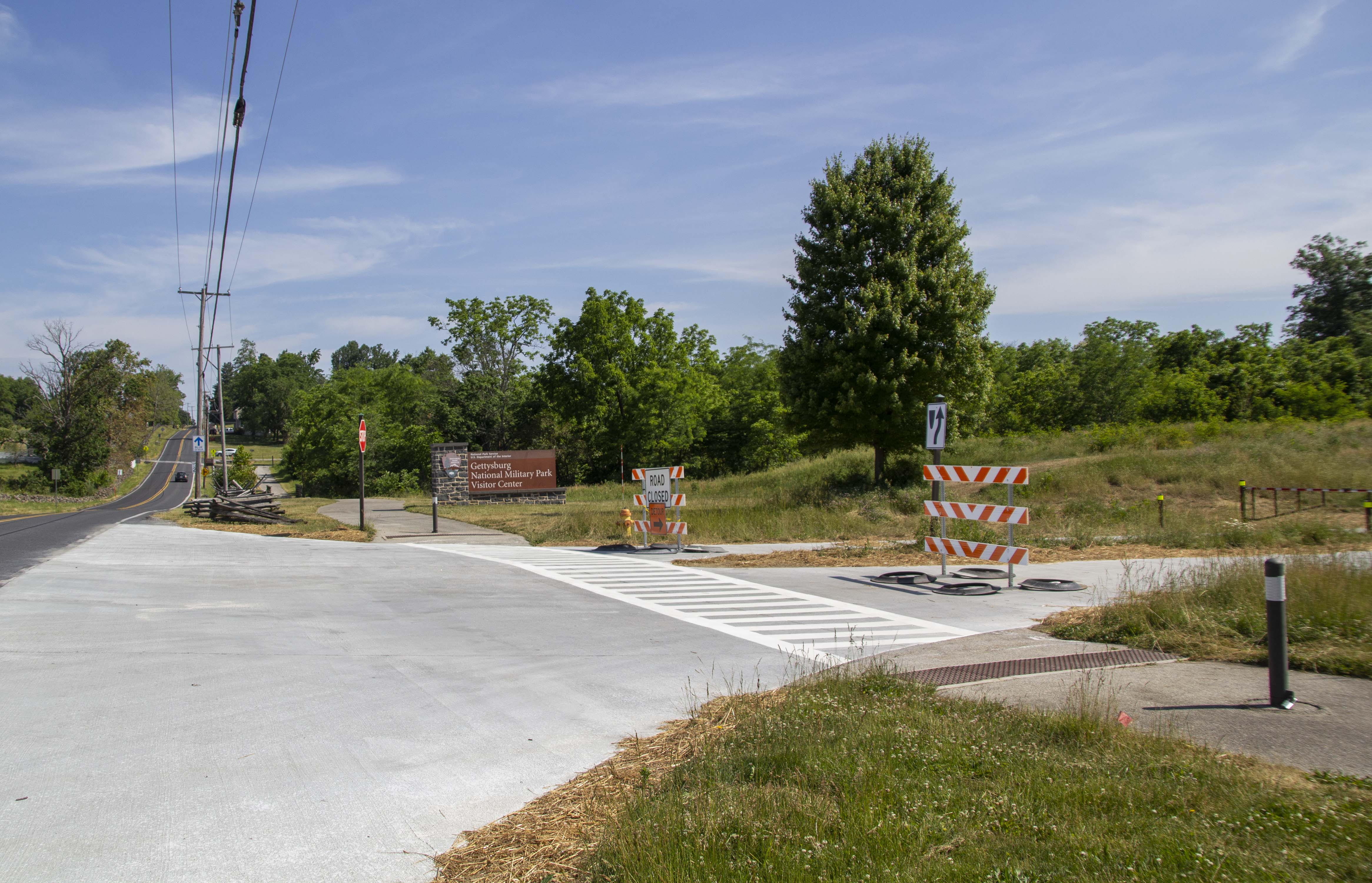 The Musuem and Visitor Center Drive at Taneytown Road will open on June 8, 2023. Photo taken from the intersection of two roads. To the left and away from the camera is a black asphalt road and the to the right is a white concrete road. There is a sidewalk running parallel to the asphalt road with white crosswalk stripes.