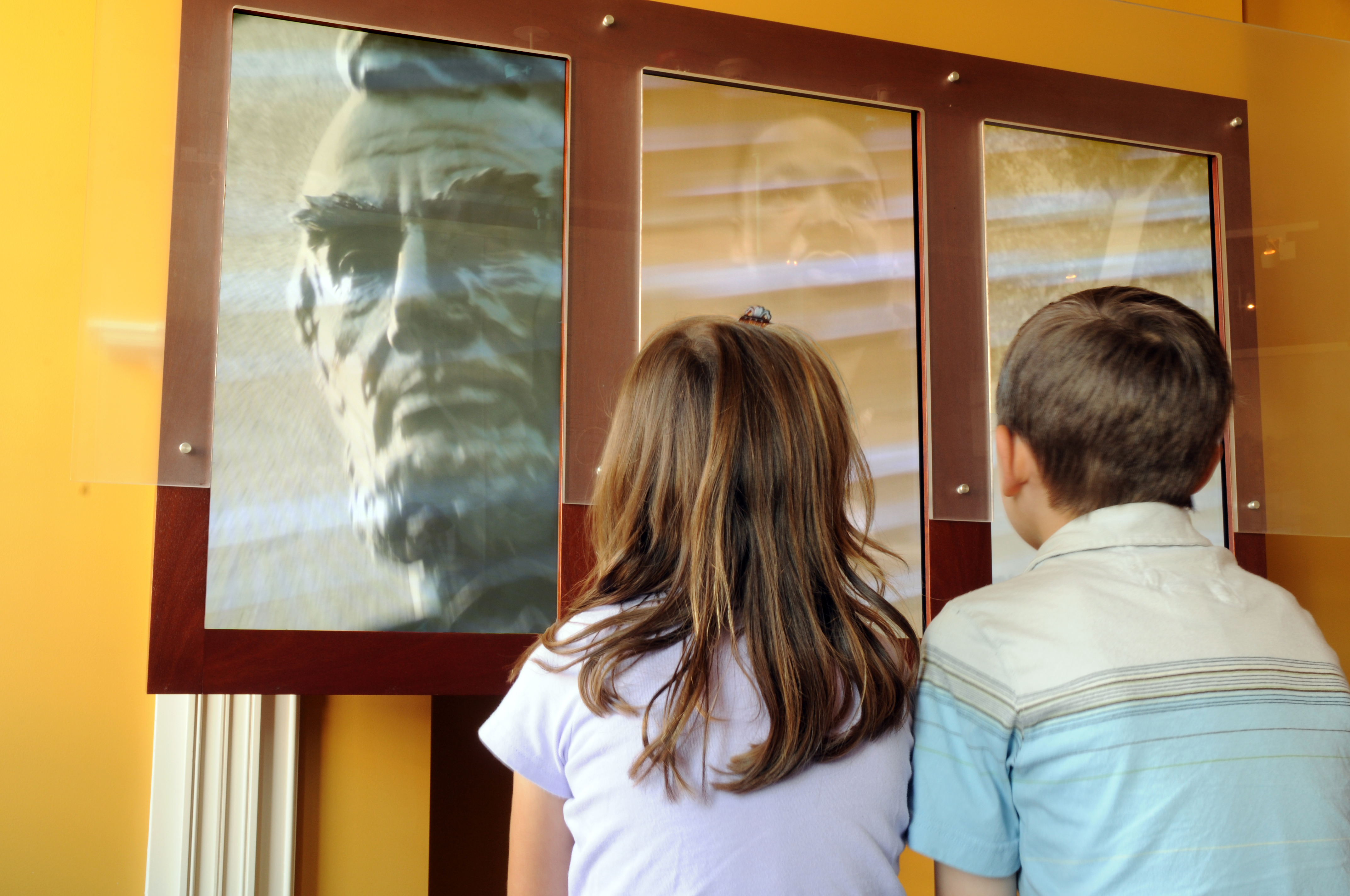 Two children, a girl on the left and a boy on the right, sit in front of a screen in a museum watching a video about Abraham Lincoln.