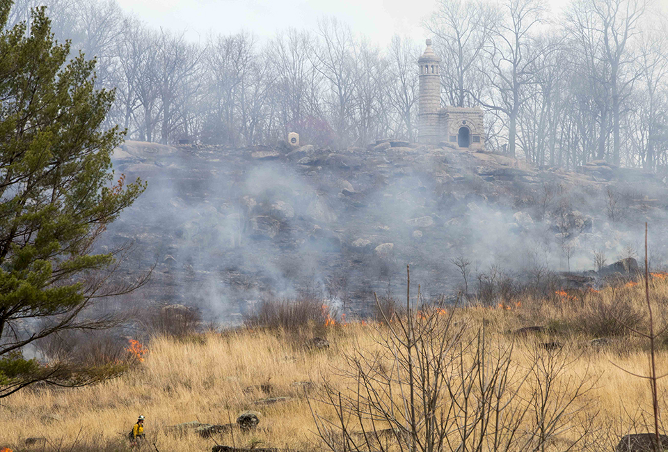 A prescribed fire moves along a yellow grass covered hill from left to right. The flames are small and the fire creates black scene behind it. A large white granite monument stands at the top of the hill in front of a line of trees.