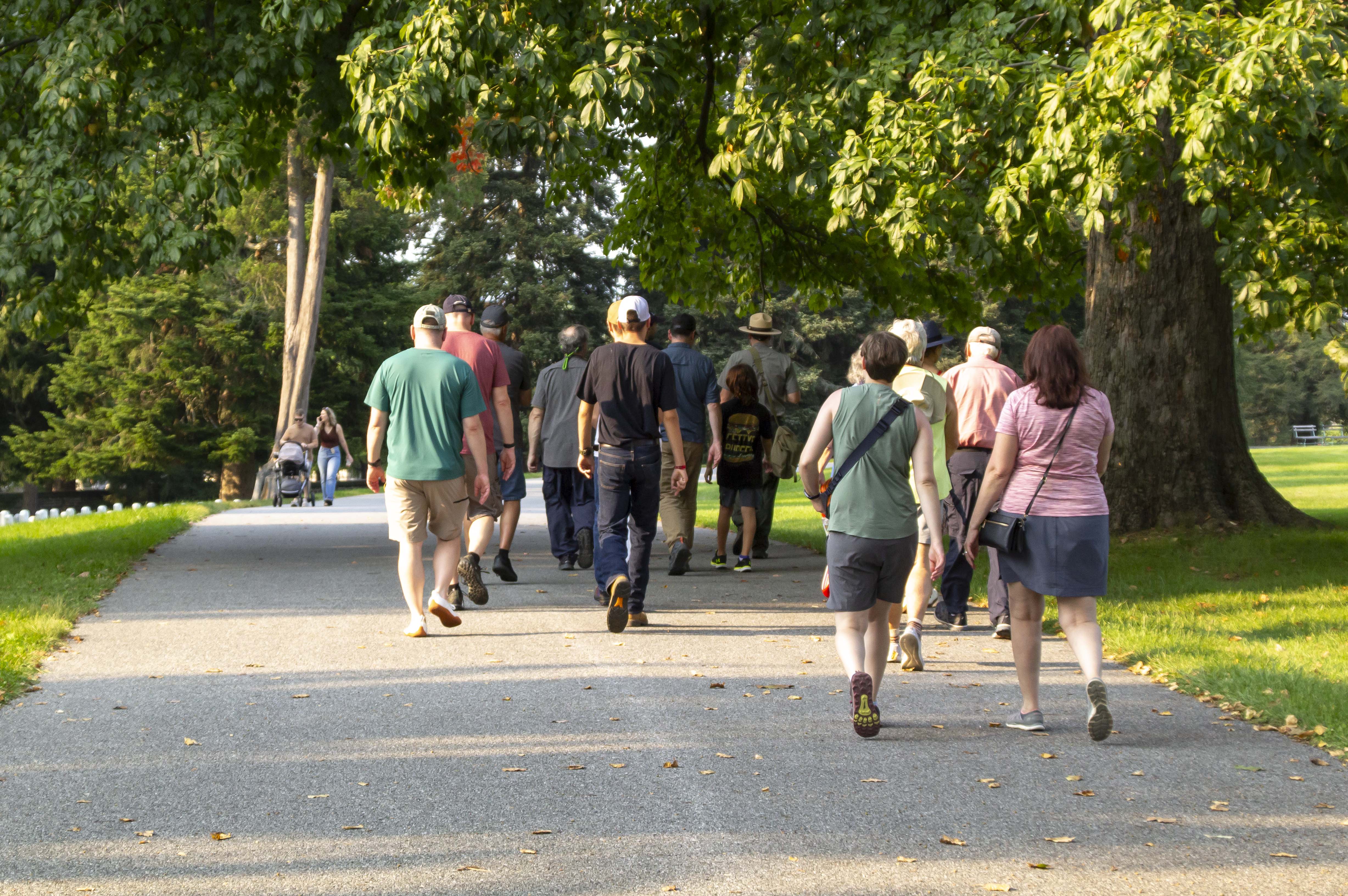 A group of visitors follow a park ranger in Gettysburg National Cemetery. A group of visitors walk away from the camera following a park ranger along a wide paved path into a cemetery.