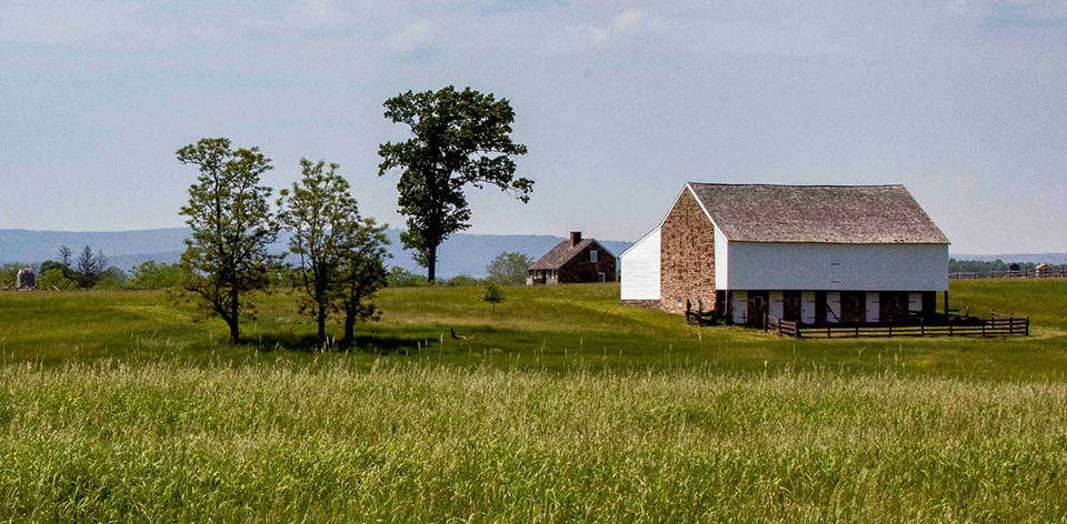 The McPherson barn, made of stone and white painted wood siding, sits on a slight ridge set among green field grasses on the right of picture. There are a few trees in the middle and another small structure in the distance.