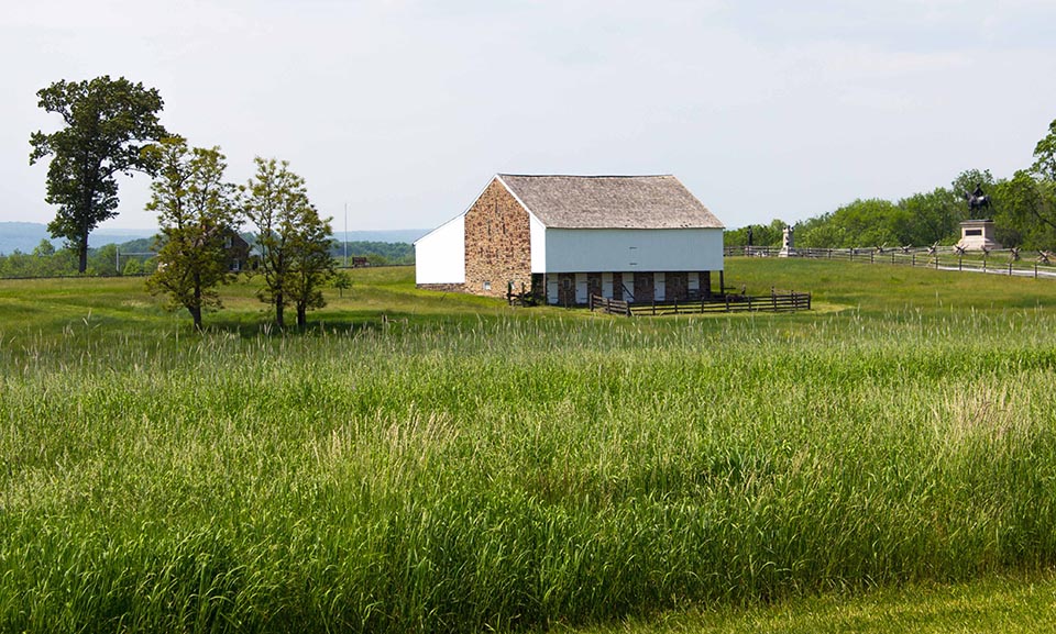 The McPherson Barn. A stone and white siding clad barn stands in the middle of a green field. A few trees stand on the left and a line of trees stands on the right. Two monuments are in the distance on the right.