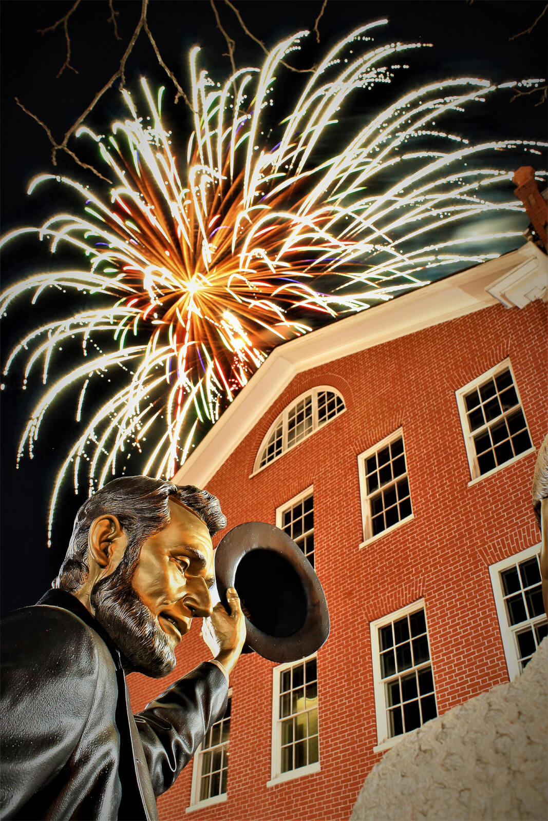 Fireworks over the David Wills house. A statue of Abrahm Lincoln stands in front of a three-story red brick house with cream color shutters. In the sky above the building are bright fireworks.