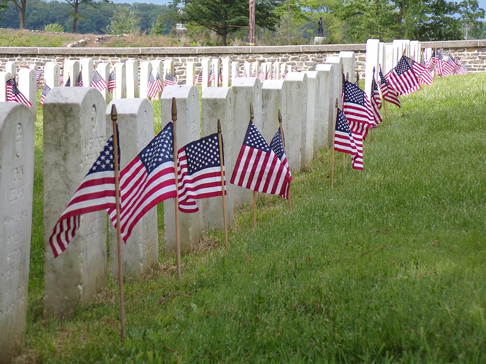 A row of white marble headstones form a line from left to right. Green grass lies in front of the headstones to the right of the picture and small American flags have been placed in front of each headstone.