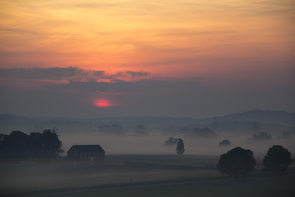 The sun rises brightly over the a foggy field. A barn is on the left and a few trees are in the center. In the distance to the right is the domed Pennsylvania monument.