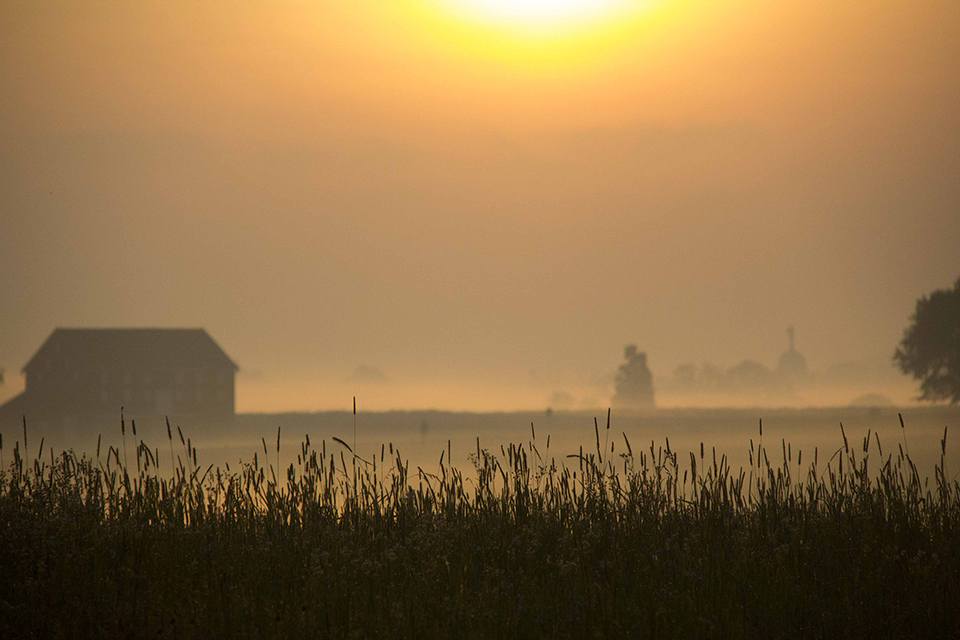 The sun rises brightly over the a foggy field. A barn is on the left and a few trees are in the center. In the distance to the right is the domed Pennsylvania monument.