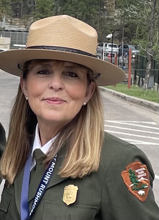 A Caucasian woman with shoulder length blond hair wears a tan broad brimmed hat, a white collared shirt, a green tie, and a green sweater. The sweater has a brown patch on the left shoulder and a gold badge over her heart. She also wears a blue lanyard.