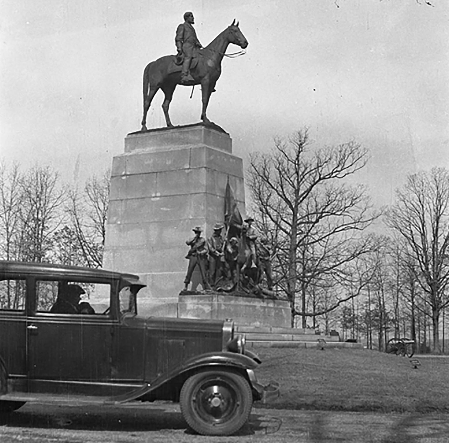 A black and white photo of a car from the early 1900s drives in front of a large monument of a soldier on a horse, on a granite pedestal, with a group of soldiers at the right bottom of the pedestal.