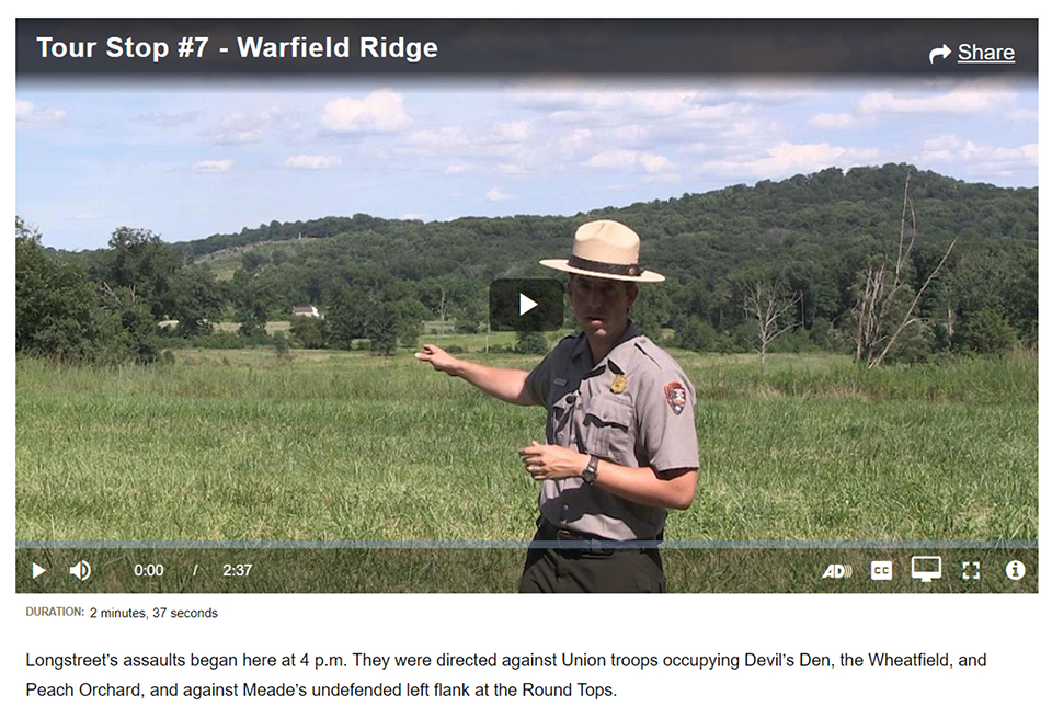 A screen shot of Virtual Tour stop seven. A park ranger, standing in a field, points off in the distance. Two tree covered hills are in the distance.