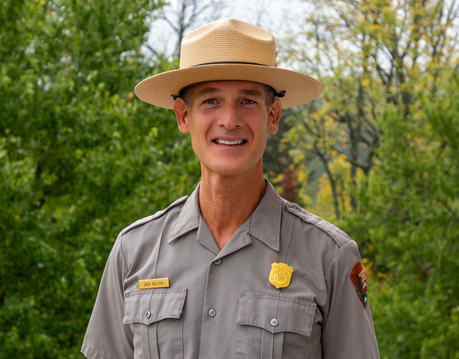 Jason_10-3-24_0002 A white male stands for a portrait. He is wearing a grey button-down uniform shirt with a patch on the left sleeve, a gold badge over his left chest, and a gold name bar over his right chest. He is wearing a flat trooper hat.