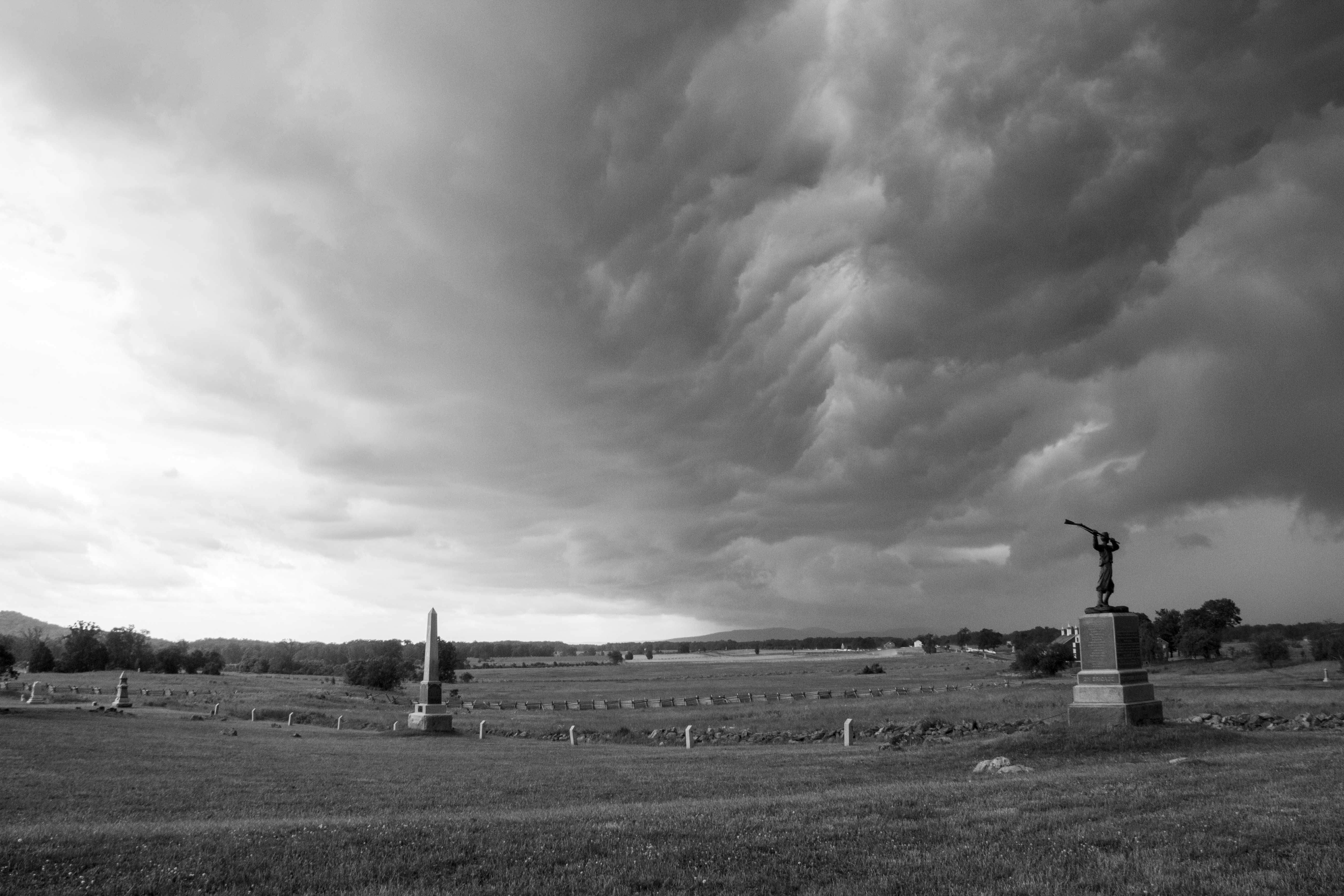 A black and white image with dark storm clouds rolling in over an open field with two monuments in the foreground and a barn in the background.