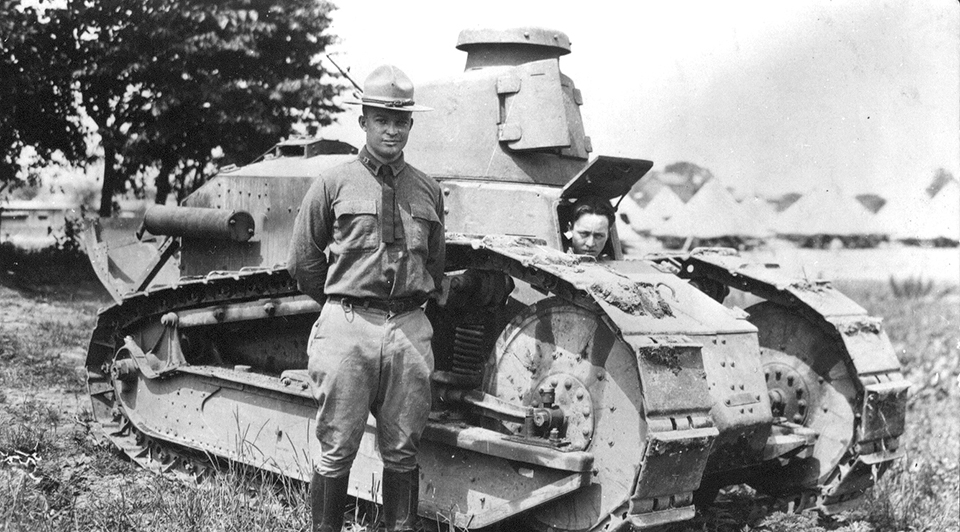 Captain Dwight D. Eisenhower stands in front of a tank in Gettysburg at Camp Colt. A black and white photograph of a young Dwight D. Eisenhower in a World War One uniform standing in front of a tank with a few trees and tents in the background.