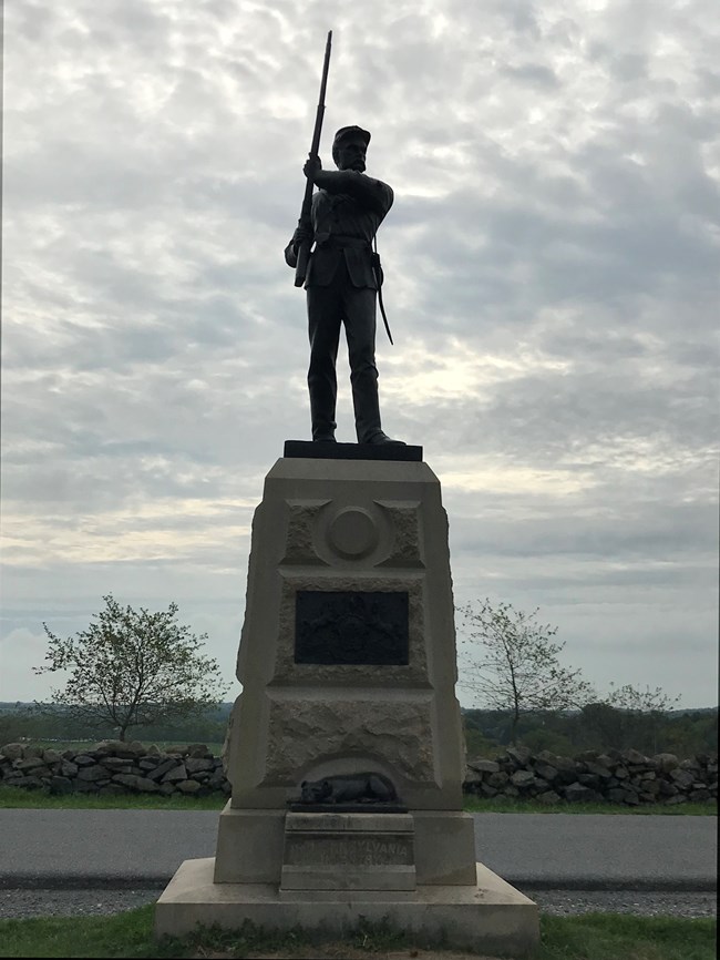 A granite monument to the 11th Pennsylvania featuring a bronze, life size statue of a soldier holding a musket, as well as a small terrier dog at monument's base