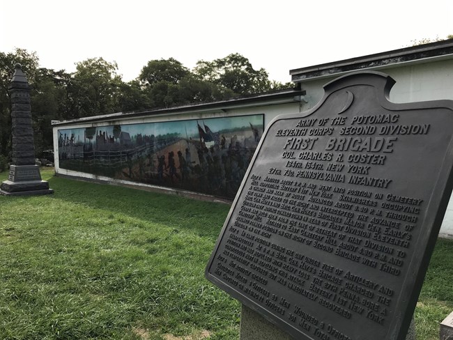 A large bronze War Department tablet appears in foreground with several granite monuments in the rear; a large painted mural depicting Civil War battle action is painted onto the wall of a neighboring building