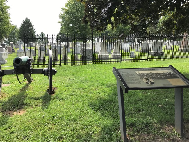 A cannon stands in the foreground, behind which is iron fence. On other side of iron fence stands numerous tombstones. A wayside panel on right is titled "The Gettysburg Address" and features a photograph of Abraham Lincoln