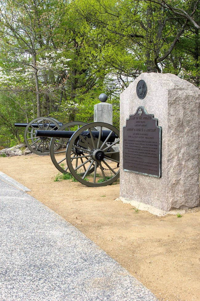 A pair of artillery pieces stand in a dirt area with a stone monument