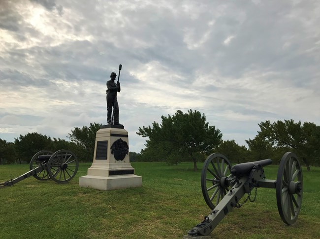 Iron cannons stand on either side of a granite monument with a bronze statue of an artillery soldier on top; in the background is a grove of young peach trees