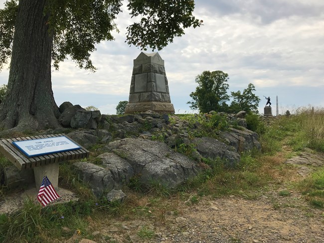 A stone wall extends along a ridgeline, behind which stands a number of granite and bronze monuments; a tree stands to the left of the stone wall and a sign reads "The Angle"