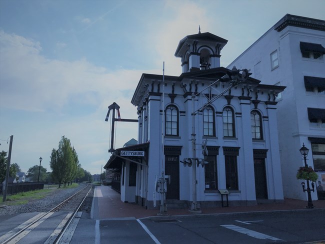 Gray Two Story Brick Train Station Building with observation platform on top and four green doors in front; railroad tracks are on left