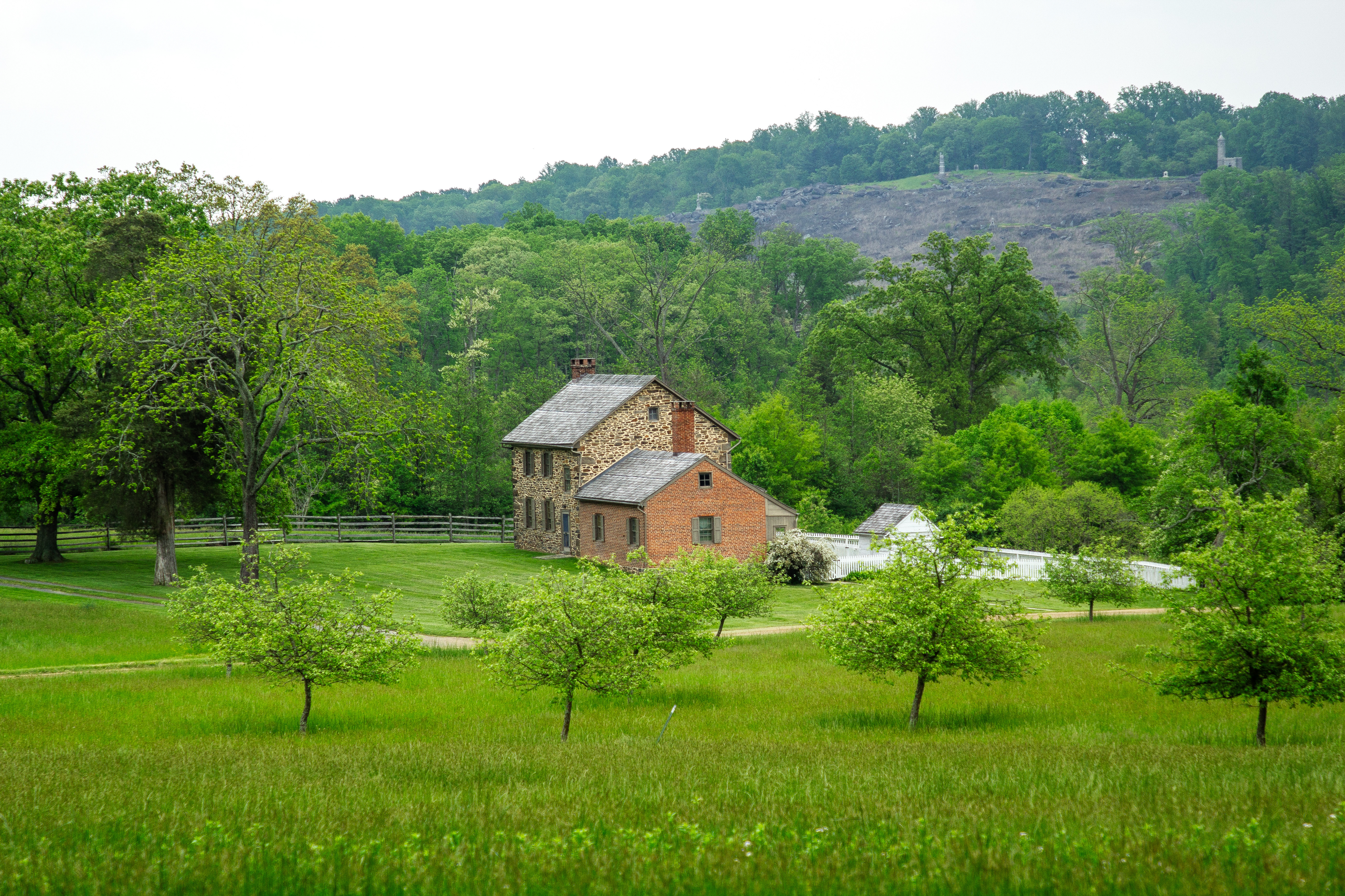 Stay in a historic home on the Gettysburg battlefield during your next visit! A stone and red brick house on grassy property with orchard trees sits in front of a heavily wooded area and a hill is in the distance.