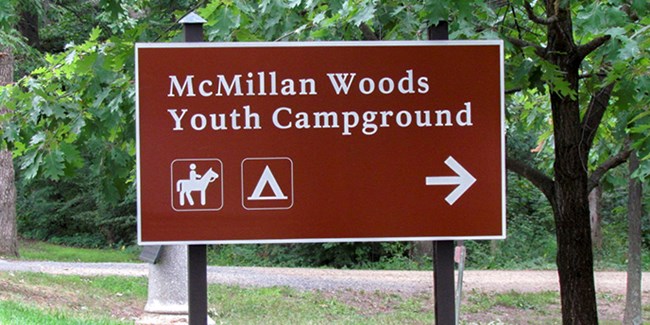 A brown sign with white letters that reads McMillan Woods Youth Campground.