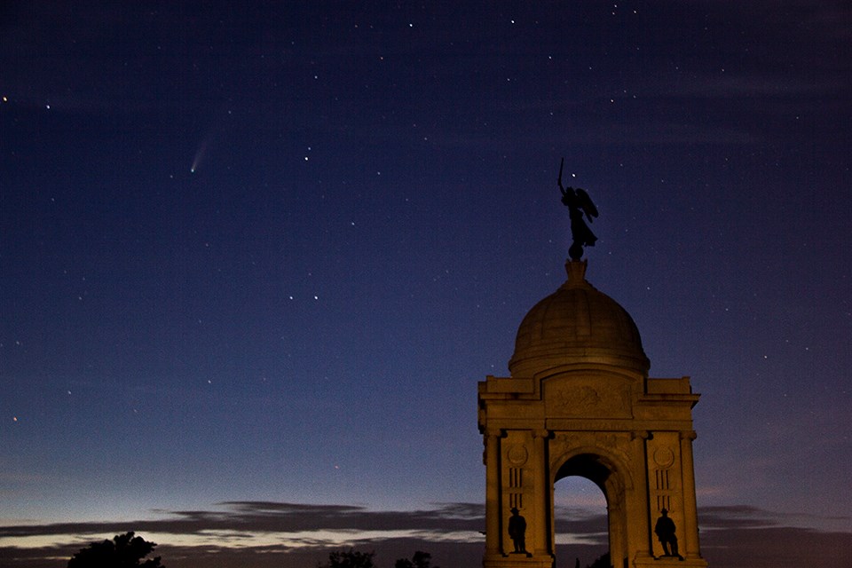 The Pennsylvania Memorial, a large domed monument with a statue at the top, is seen at early evening with a starry sky and a comet in the upper left corner.