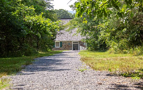 A gravel path is lined with trees and there is a small stone building with green shutters in the distance.