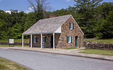 A small stone building with green shutters sits next to a road. A stone wall surrounds it and a gravel path leads from the building to the right through the trees.