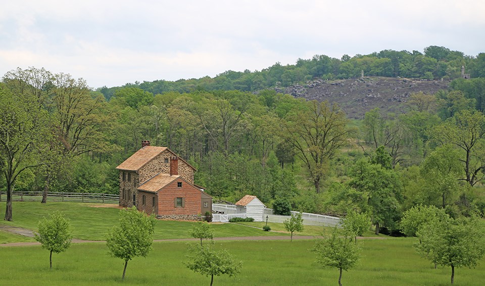 The brick and stone Bushman house is nestled among a green field, orchards, and woods. In the distance is the slope of Little Round Top.