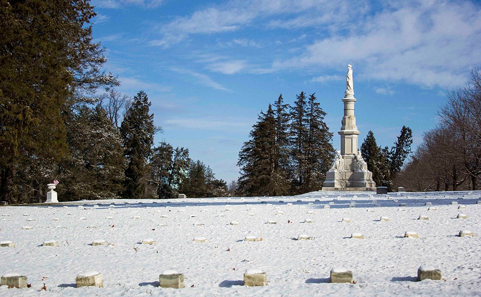 Snow covers the ground in the National Cemetery. The Soldiers' National Monument stands against a blue sky.
