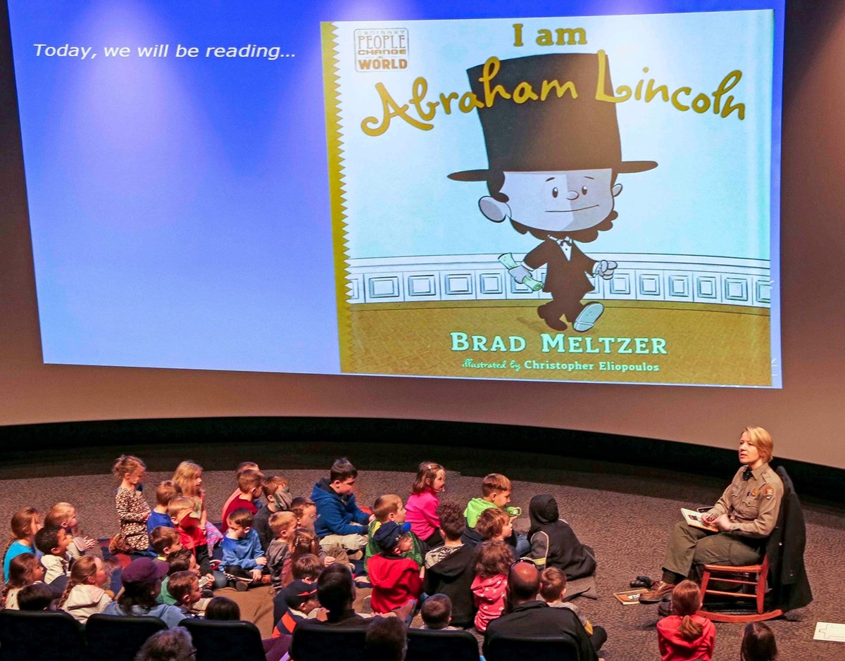 A seated Park Ranger reads a children's book about Abraham Lincoln in front of a large number of children