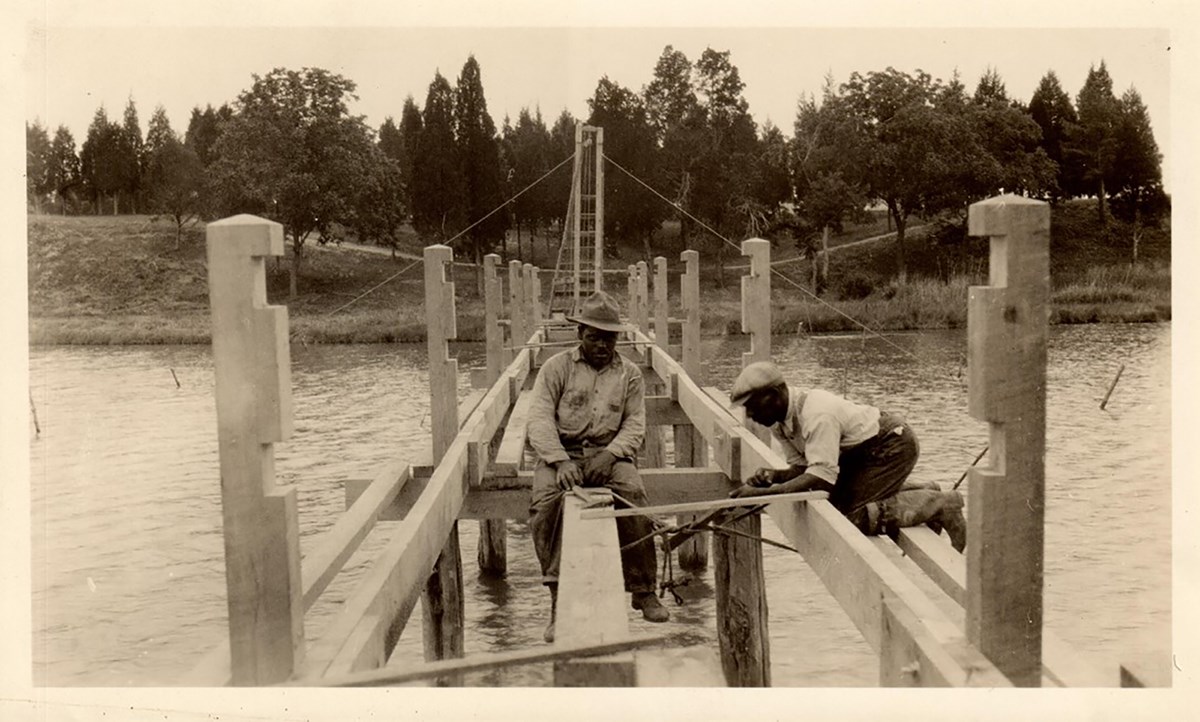 2 black men building a bridge over water in the park.
