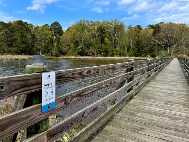 bridge above water, a plastic holder for a phone sits a top of the bridge railing. Below is a sign for the Chronolog station. Overlooking a shore line and treese