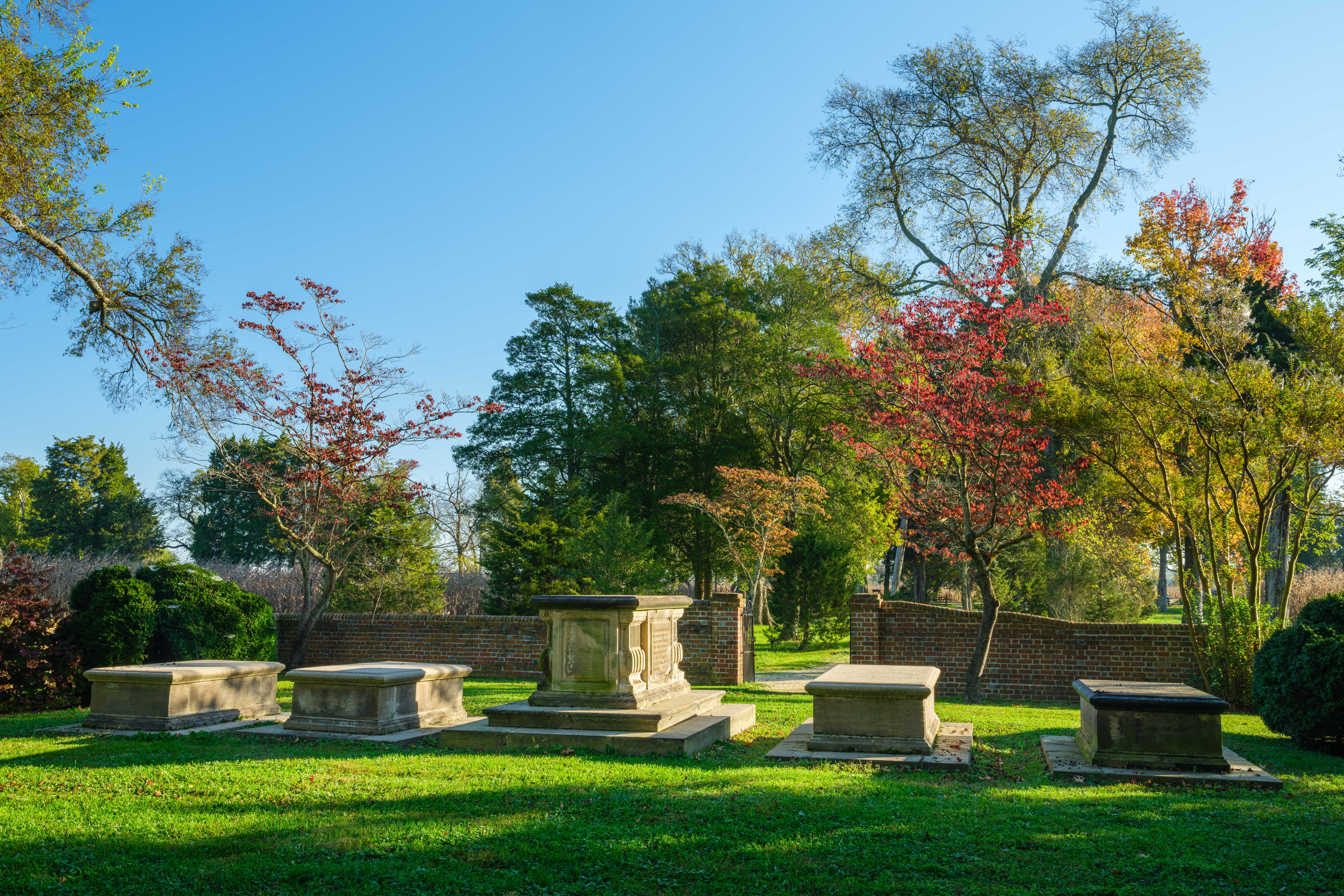 Several gravestones lay in a grass field surrounded by a brick wall with trees and a blue sky.