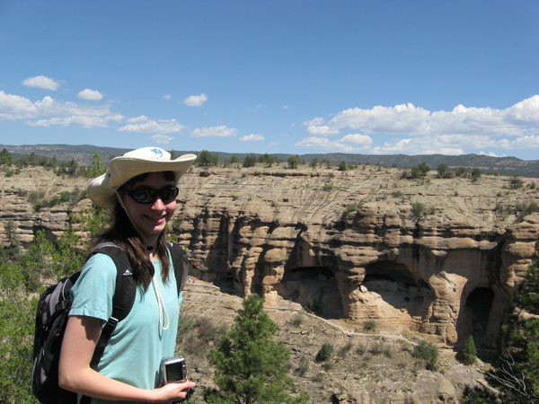 A student intern at Gila Cliff Dwellings National Monument smiles over a view of the cliff dwellings from the opposite side of the canyon.