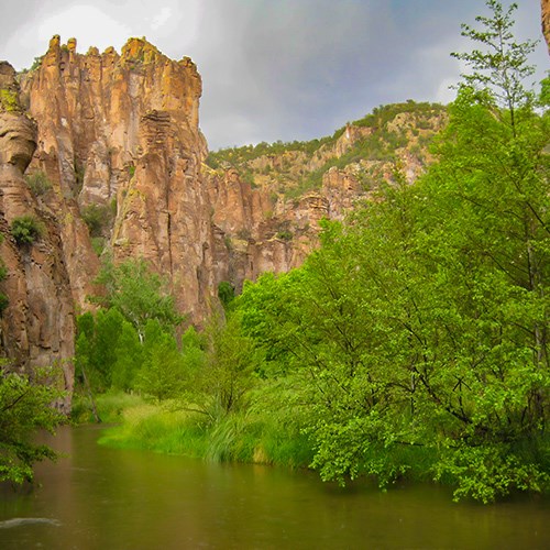 A river flows with lush green vegetation next to it and high cliff formations in the background.l