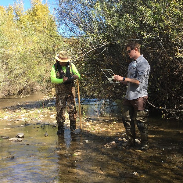 Two men stand in a shallow stream. One writes on a clipboard.