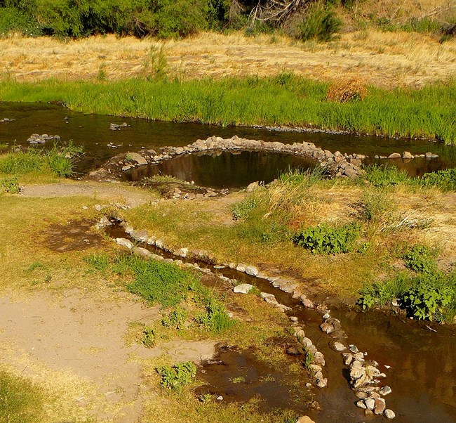 A rock-lined channel winds out to a rock-lined pool built in the middle of a river surrounded by bright green grass.