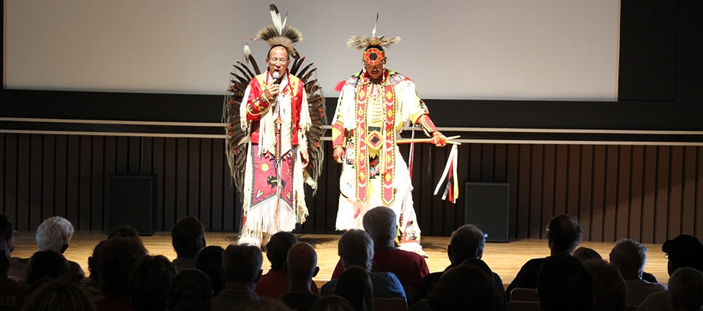 audience members in silhouette as two tribal members in ceremonial dress stand on stage audience members in silhouette as two tribal members in ceremonial dress stand on stage