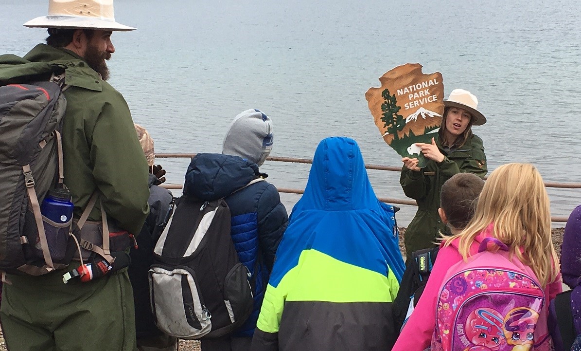 Ranger watches over line of students by a lake while another ranger holds up NPS arrowhead and points to the bison Ranger watches over line of students by a lake while another ranger holds up NPS arrowhead and points to the bison